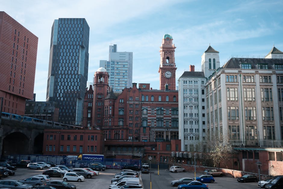 A street scene in Manchester showcasing a variety of building facades, including a classic red brick building with multiple windows and a decorative cornice, surrounded by modern glass skyscrapers wit