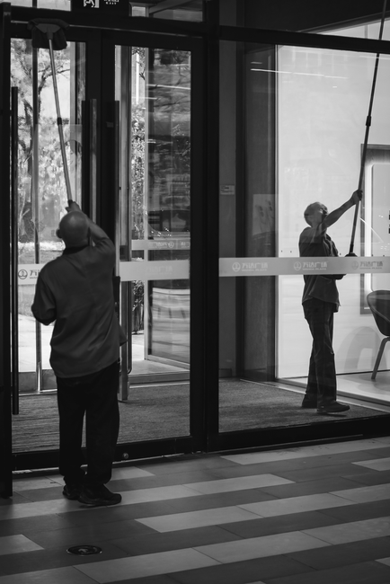 A female commercial cleaner wearing a grey t-shirt and bright orange apron is seen in a well-lit, modern kitchen space. She is holding a blue cleaning bucket placed on a white countertop. The surface